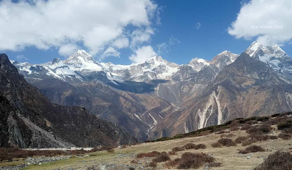 A wide panoramic view of jagged snow capped Himalayan peaks towering over a rugged mountain valley under a bright cloudy sky