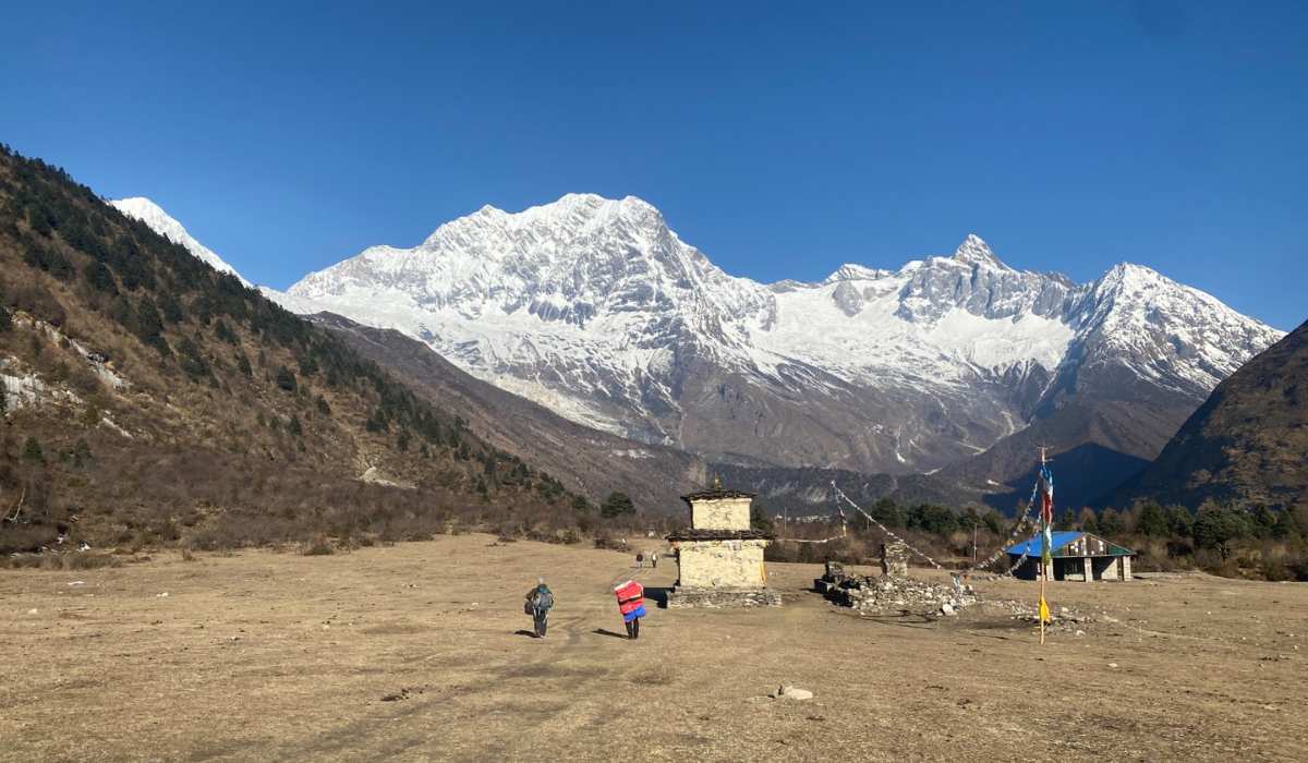 Two trekkers walk toward a stone chorten in a wide Himalayan valley framed by towering snow capped peaks and a clear blue sky