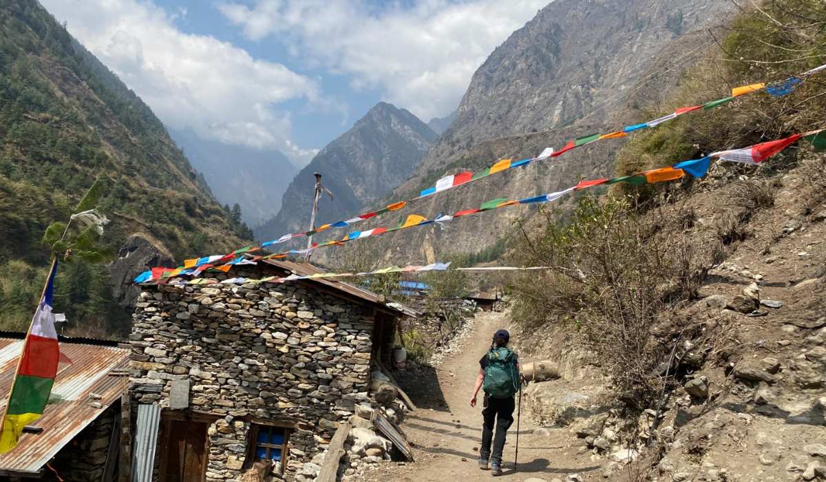 our guest walks down a dirt path past a stone house decorated with colorful prayer flags leading into a deep mountain valley